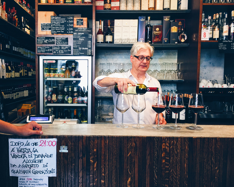 A white haired man standing behind a shop counter pours red wine in five glasses