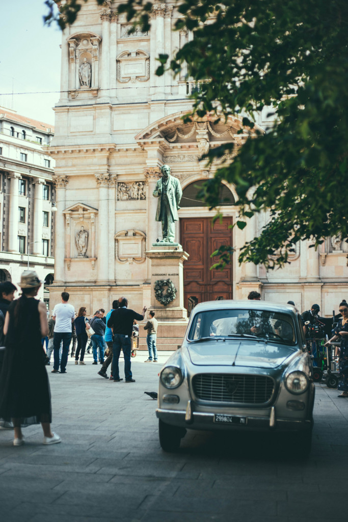 People walk past a vintage car parked in an Italian piazza while doing lo struscio